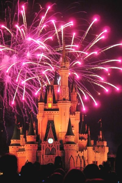 Cinderella's Castle at Magic Kingdom, lit up at night with fireworks overhead.